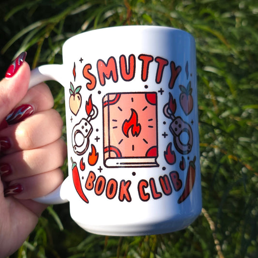 Person holding a mug with 'Smutty Book Club' design against a green leafy background